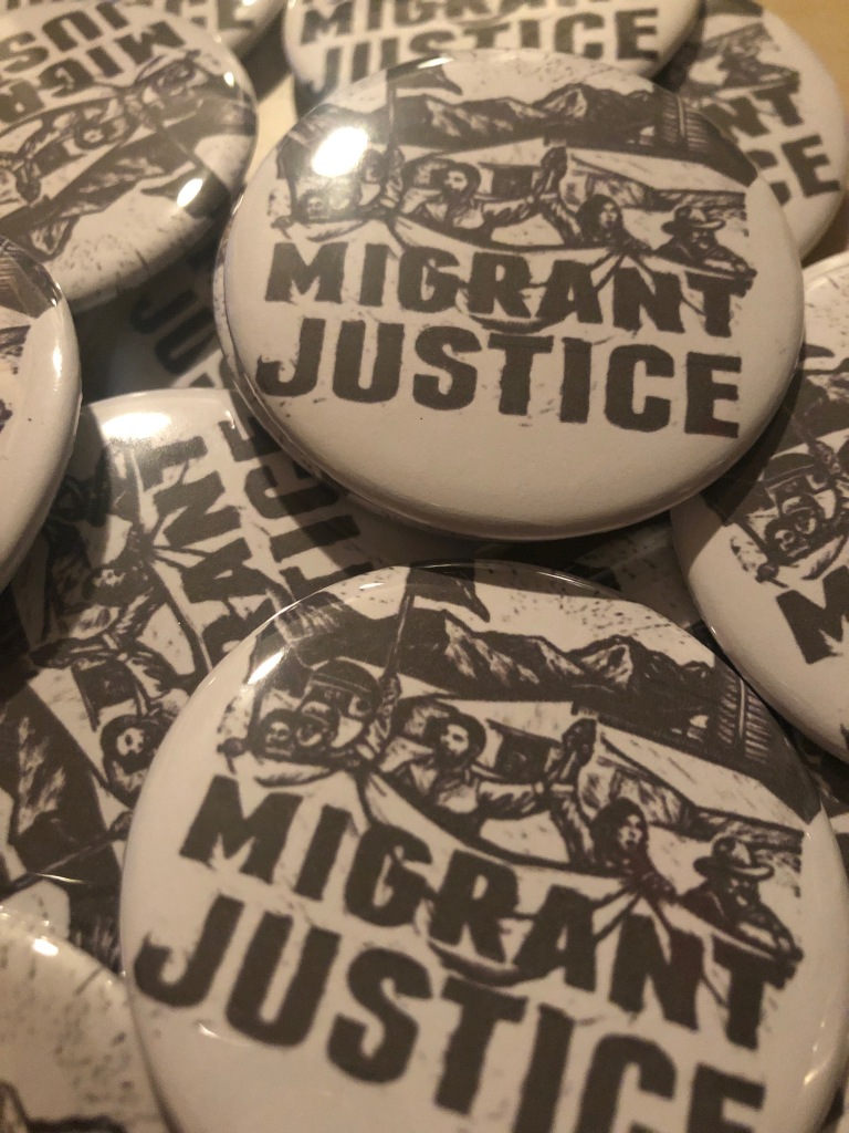 Close-up photo of buttons created by BakaGirlJewelry for the event. Buttons say Migrant Justice in all-caps, with drawing of people with arms raised and mouths open.
